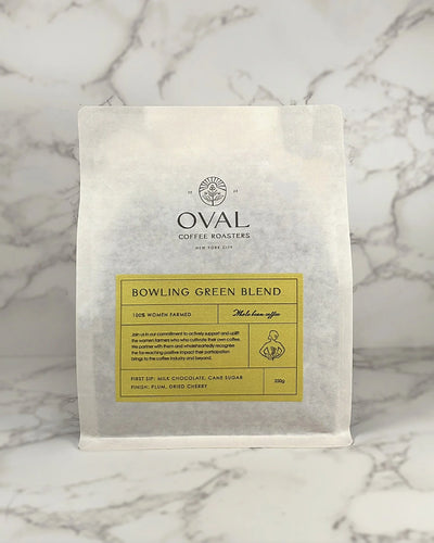 Oval Coffee Roasters Bowling Green Women-Grown light roast 8.82 oz whole-bean coffee bag with a yellow label, woman holding a coffee branch illustration, and black Oval logo on gray-veined marble.