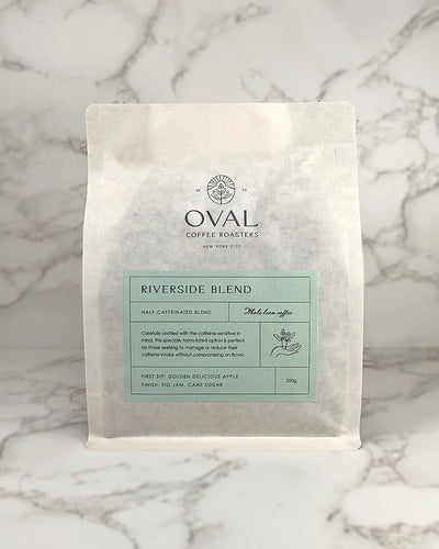 Oval Coffee Roasters Riverside Half-Caf 8.82 oz whole-bean coffee bag with a pale green label, coffee-branch hand illustration, and black Oval logo on gray-veined marble; a light-to-medium roast.