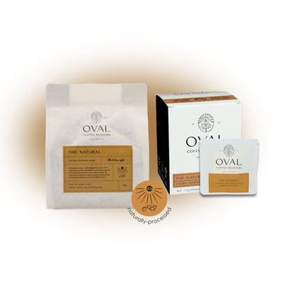 Oval Coffee Roasters The Natural whole-bean bag, eight-count single-serve pour-over box and individual drip-bag sleeve with orange labels beside a circular Natural Process badge.