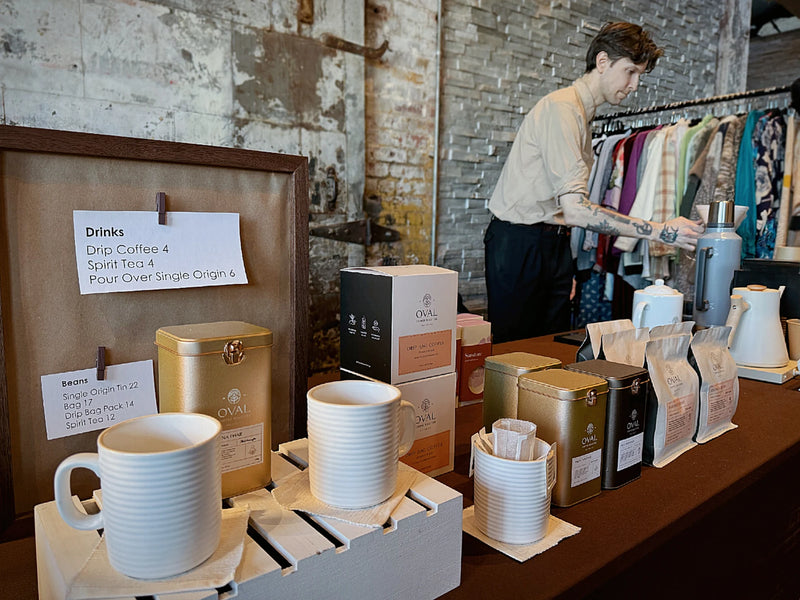 Christopher Alspach preparing coffee behind an Oval Coffee Roasters market table in Brooklyn, with gold coffee tins, whole-bean bags, drip-bag boxes and white cups at the Greenpointers Valentine’s Day Market.
