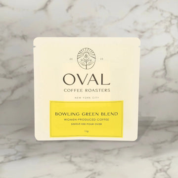Front of an Oval Coffee Roasters Bowling Green Women-Grown light roast coffee 0.39 oz single-serve pour-over sleeve, with a yellow label and black Oval logo against gray-veined marble.
