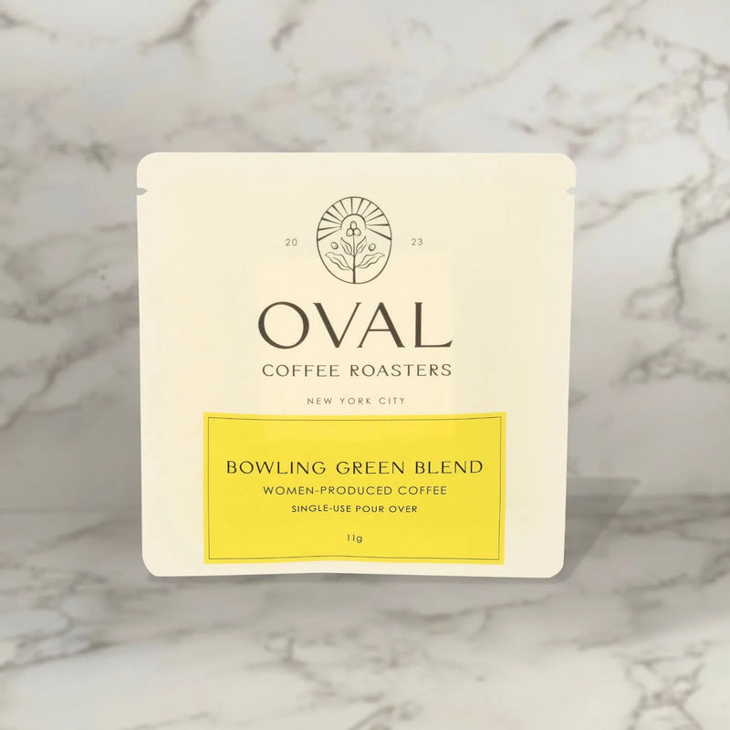 Front of an Oval Coffee Roasters Bowling Green Women-Grown light roast coffee 0.39 oz single-serve pour-over sleeve, with a yellow label and black Oval logo against gray-veined marble.