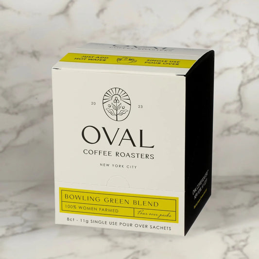 Oval Coffee Roasters Bowling Green Women-Grown light roast coffee eight-pack box, with a yellow label and ‘Just add hot water’ strip, containing eight 0.39 oz single-serve pour-over sachets against gray-veined marble.