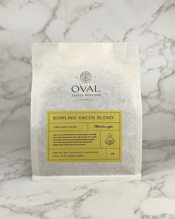 Oval Coffee Roasters Bowling Green Women-Grown light roast 8.82 oz whole-bean coffee bag with a yellow label, woman holding a coffee branch illustration, and black Oval logo on gray-veined marble.