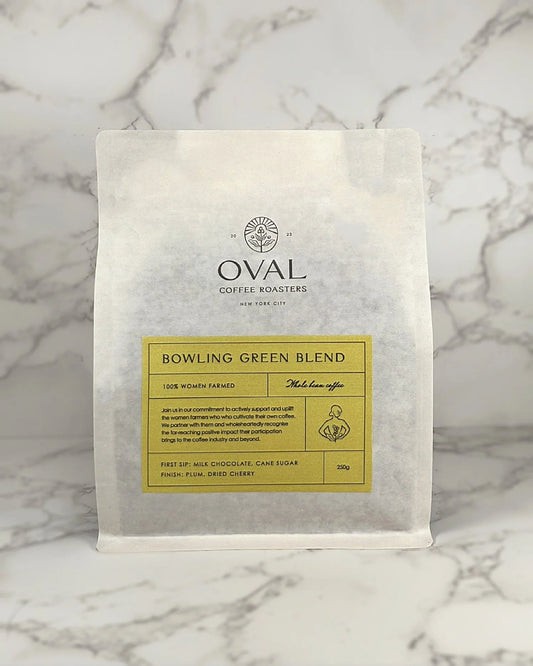 Oval Coffee Roasters Bowling Green Women-Grown light roast 8.82 oz whole-bean coffee bag with a yellow label, woman holding a coffee branch illustration, and black Oval logo on gray-veined marble.