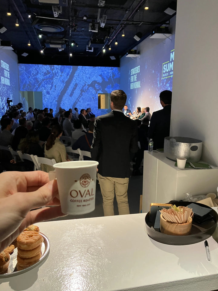 A hand holds an Oval Coffee Roasters paper cup beside pastries at Lume Studios in Tribeca, New York City, with seated attendees and blue wall projections behind the coffee counter.