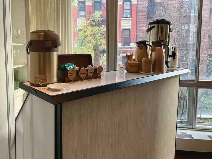 Oval Coffee Roasters catering counter beside windows at a Midtown Manhattan event, with a stainless-steel coffee urn, gold thermal carafes, paper cups and labeled tea selections overlooking brick buildings.