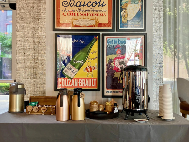 Oval Coffee Roasters coffee catering station at One Manhattan Square, New York City, with a stainless-steel urn, gold thermal carafes, condiments and stacked paper cups beneath framed vintage posters.