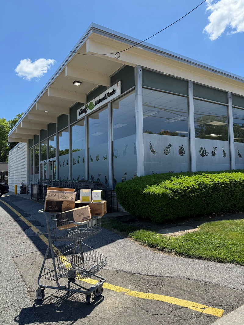 Wholesale delivery of Oval Coffee Roasters cases and coffee packages in a shopping cart outside Green’s Natural Foods in Eastchester, Westchester County, New York, beneath a blue sky.