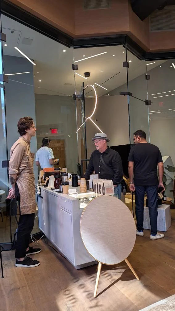 Guests gather around an Oval Coffee Roasters pop-up counter at Bang & Olufsen in SoHo, New York City, with coffee bags, brewers and stacked cups beside a large circular speaker on wooden legs.
