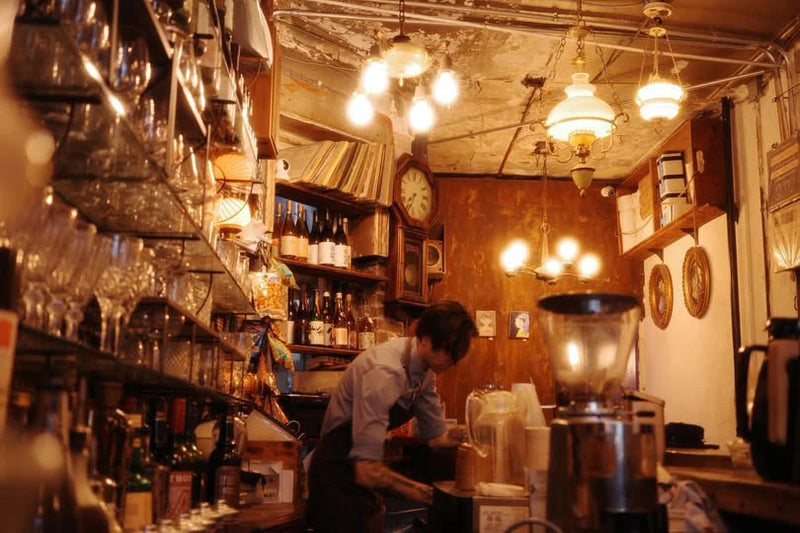 Christopher Alspach working behind the Oval coffee counter at Hard To Explain in New York City’s East Village, surrounded by glassware, a coffee grinder and glowing vintage pendant lights.