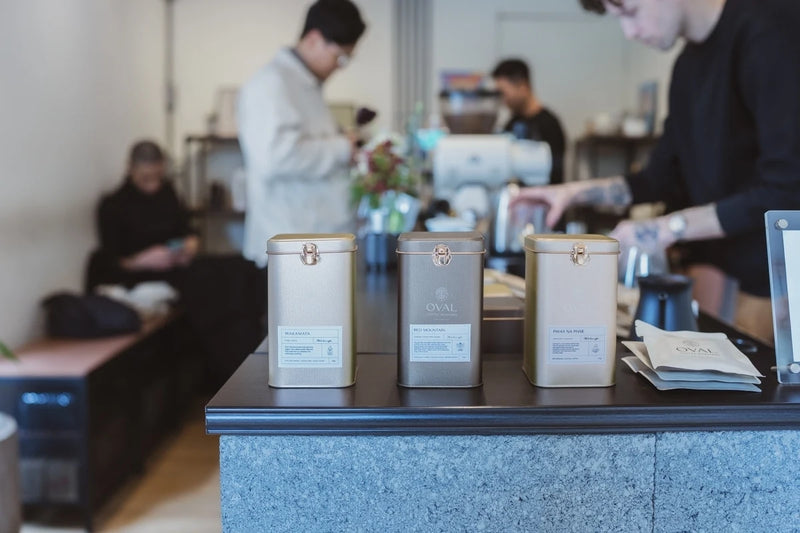 Three rectangular Oval coffee tins lined up on the stone-fronted counter at Kikumo Coffee in Taipei, Taiwan, with Christopher Alspach preparing coffee behind them and single-serve packets to the right.