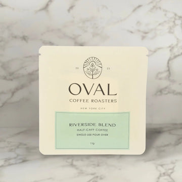 Front of an Oval Coffee Roasters Riverside Half-Caf 0.39 oz single-serve pour-over coffee sleeve, with a pale green label and black Oval logo against gray-veined marble.