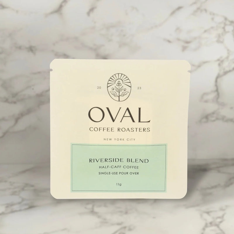 Front of an Oval Coffee Roasters Riverside Half-Caf 0.39 oz single-serve pour-over coffee sleeve, with a pale green label and black Oval logo against gray-veined marble.