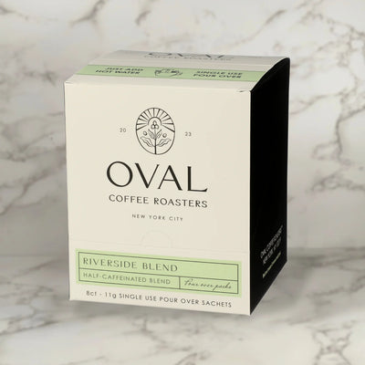 Oval Coffee Roasters Riverside Half-Caf eight-pack single-serve pour-over coffee box with a pale green label, black side panel, and “Just add hot water” strip on gray-veined marble; contains eight 11 g sachets.