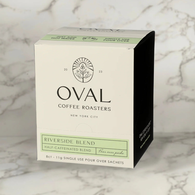 Oval Coffee Roasters Riverside Half-Caf eight-pack single-serve pour-over coffee box with a pale green label, black side panel, and “Just add hot water” strip on gray-veined marble; contains eight 0.39 oz sachets.