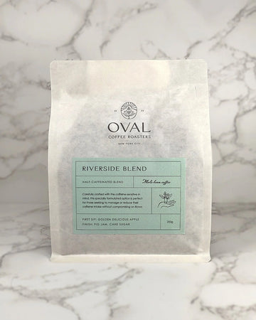 Oval Coffee Roasters Riverside Half-Caf 8.82 oz whole-bean coffee bag with a pale green label, coffee-branch hand illustration, and black Oval logo on gray-veined marble; a light-to-medium roast.
