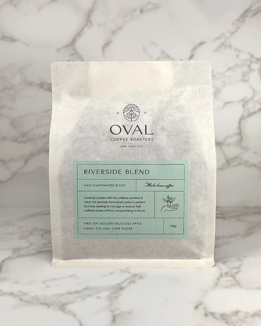 Oval Coffee Roasters Riverside Half-Caf 8.82 oz whole-bean coffee bag with a pale green label, coffee-branch hand illustration, and black Oval logo on gray-veined marble; a light-to-medium roast.