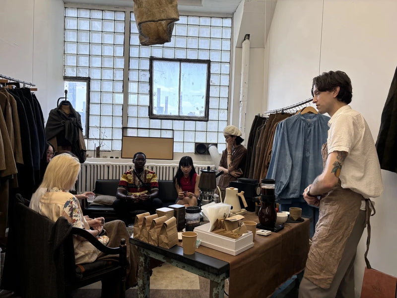 Oval Coffee Roasters pop-up coffee table at Aviva Jifei Xue’s studio in Queens, New York City, with brewers, a kettle and kraft coffee bags surrounded by seated guests, clothing racks and glass-block windows.