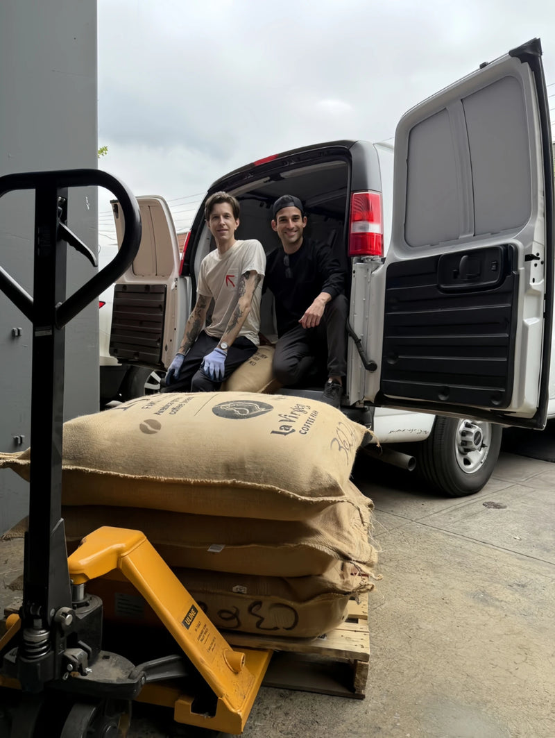 Oval Coffee Roasters co-founders Christopher Alspach and Andrew Itzkowitz sit at the open rear of a white delivery van beside burlap green-coffee sacks on a pallet and a yellow pallet jack in the Bronx, NYC.