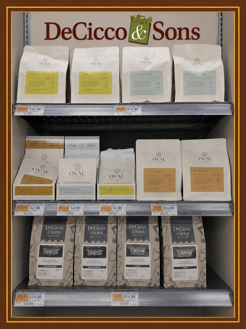 Oval Coffee Roasters retail display beneath a DeCicco & Sons sign, with Bowling Green Women-Grown and Riverside Half-Caf whole-bean bags above single-serve pour-over sleeves and The Natural bags, photographed for the Westchester and Putnam County launch.