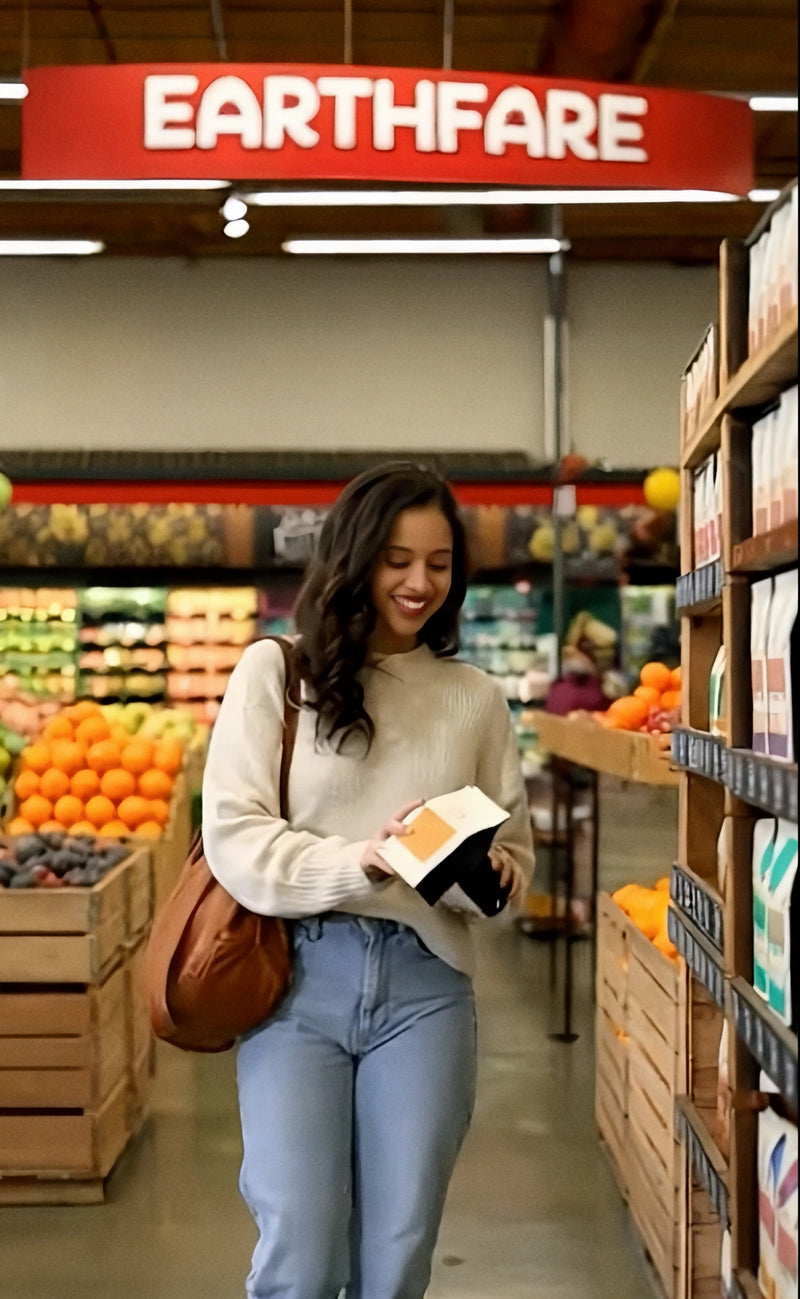 A shopper holds an orange-labeled Oval Coffee Roasters The Natural whole-bean coffee bag beside grocery shelves and produce beneath an Earth Fare sign, from the brand’s Southeastern retail launch imagery.
