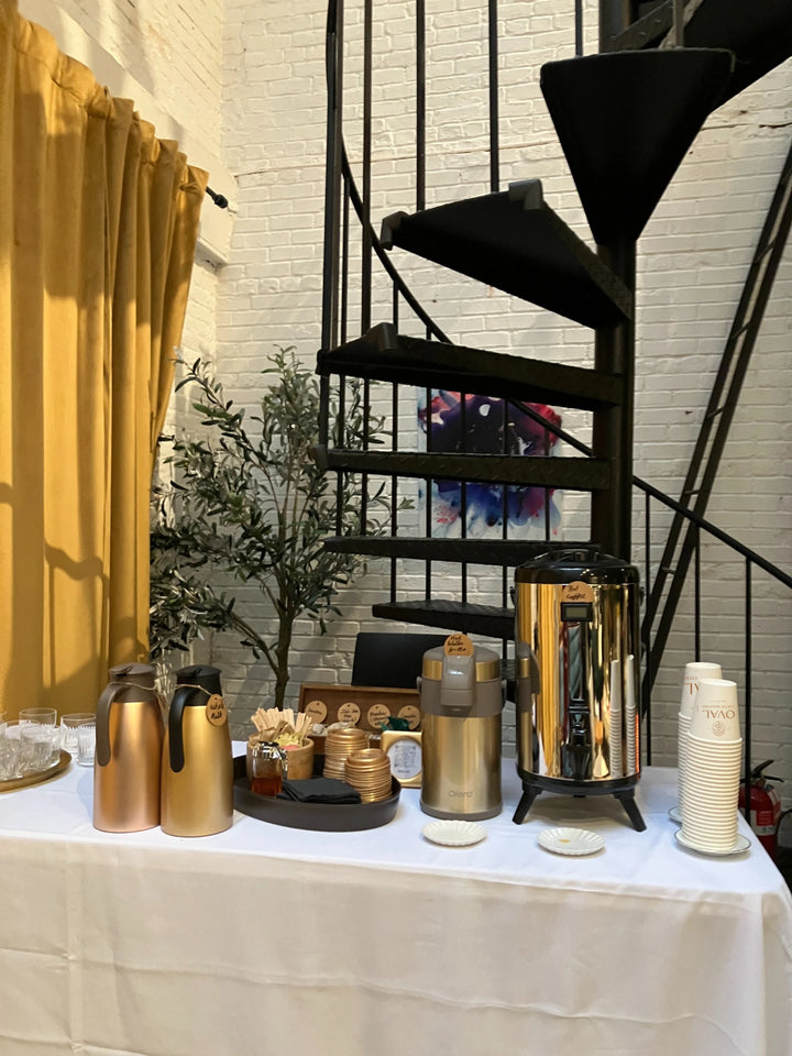 Oval Coffee Roasters event catering table beneath a black spiral staircase in New York City, with gold thermal carafes, a stainless-steel coffee urn, condiments and stacked Oval cups against a white brick wall.
