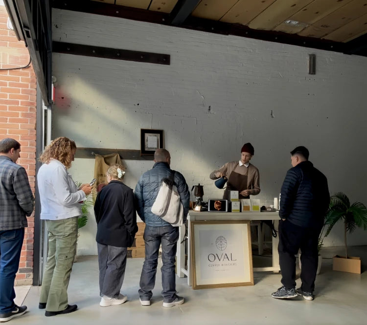 A barista pours coffee for guests at a December Oval Sundays pop-up near the Brooklyn Navy Yard, with a framed Oval Coffee Roasters sign and coffee packages beneath white brick walls and exposed ceiling beams.