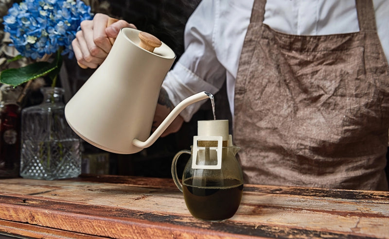 A barista wearing an Aviva Jifei Xue apron pours hot water from a white Fellow Stagg gooseneck kettle into an Oval Coffee Roasters single-serve drip filter over a glass server, beside blue flowers in NYC’s East Village.