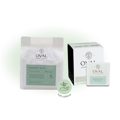 Oval Coffee Roasters Riverside Half Caff whole-bean bag, eight-count single-serve pour-over box and individual drip-bag sleeve with pale green labels beside a circular half-caffeine badge.