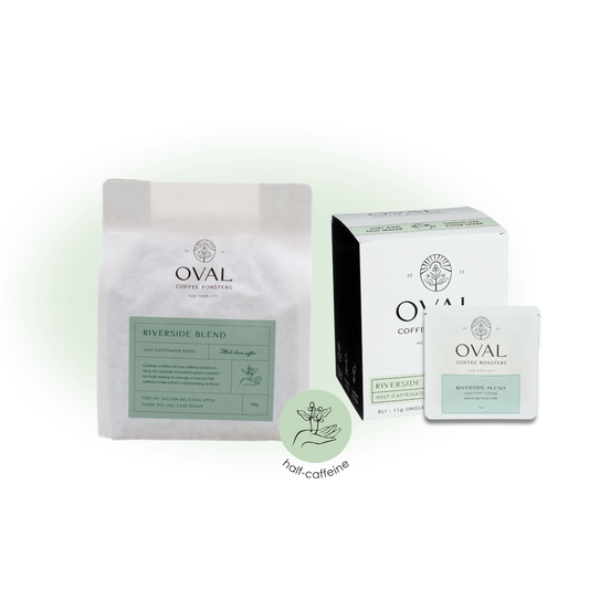 Oval Coffee Roasters Riverside Half Caff whole-bean bag, eight-count single-serve pour-over box and individual drip-bag sleeve with pale green labels beside a circular half-caffeine badge.