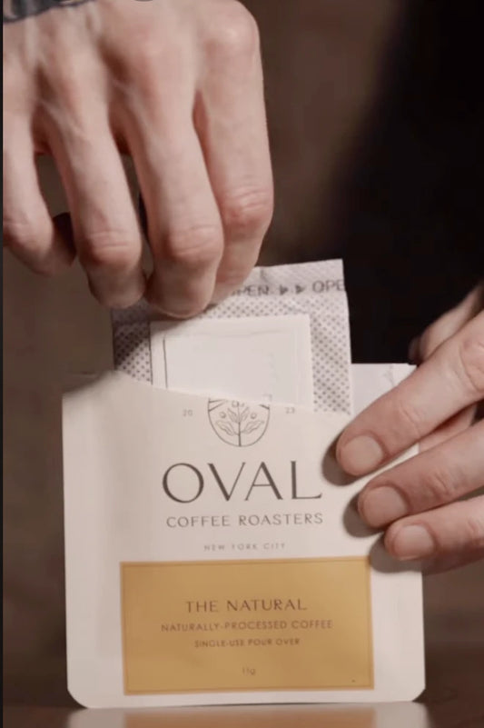 Hands lift a white drip filter from an opened Oval Coffee Roasters The Natural sachet with an orange label, demonstrating single-serve pour-over natural process coffee.