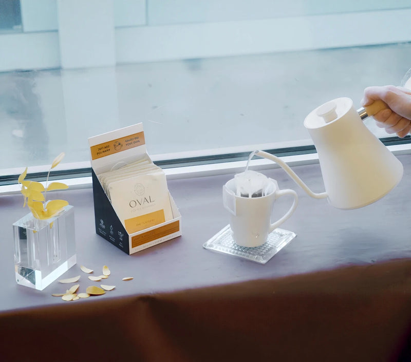A white Fellow Stagg gooseneck kettle pours hot water into an Oval Coffee Roasters drip filter over a white mug beside an open The Natural coffee caddy and yellow leaves, from the single-serve pour-over brewing tutorial.