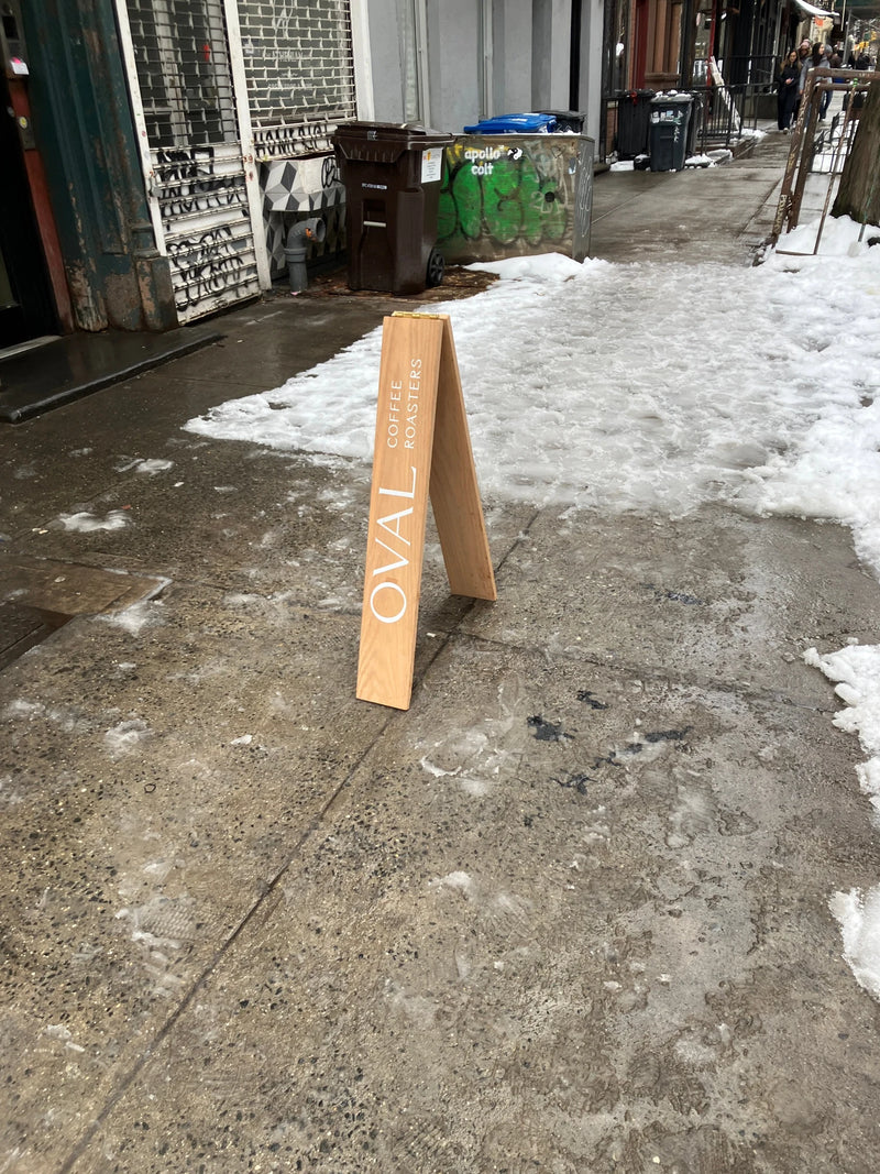 Tall wooden A-frame sign reading Oval Coffee Roasters on a wet, snow-lined sidewalk outside East Village storefronts in New York City.