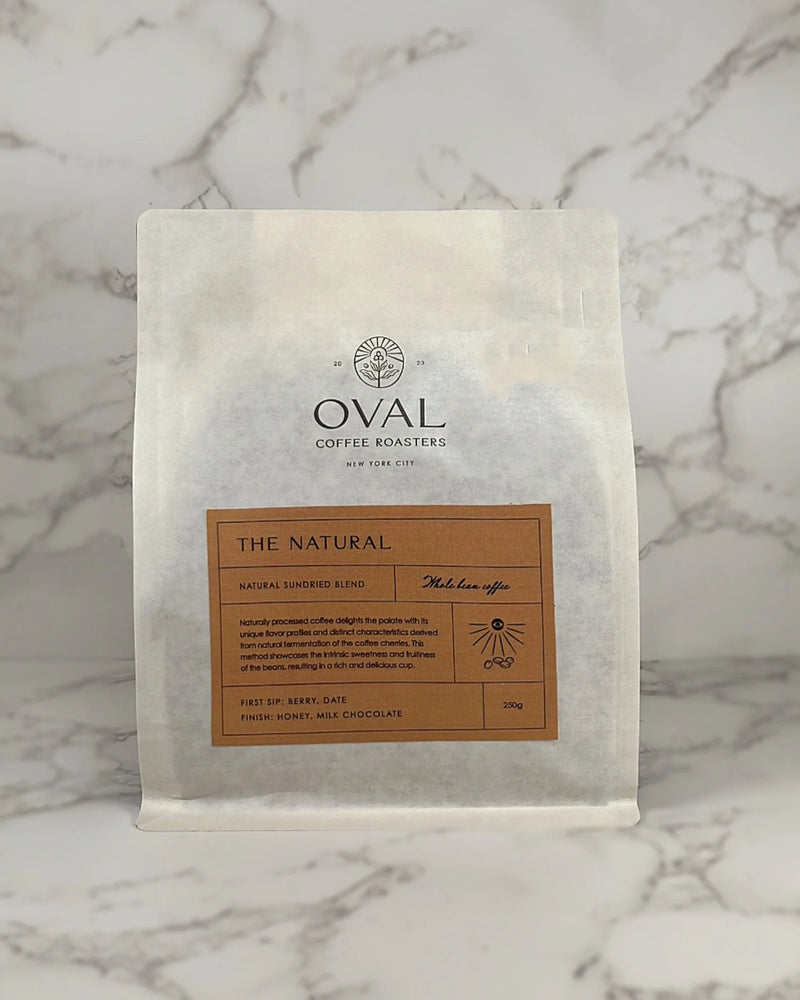 Oval Coffee Roasters The Natural 8.82 oz whole-bean natural process coffee bag with an orange label, sun-and-coffee-cherry illustration, and black Oval logo on gray-veined marble; a light-to-medium roast.
