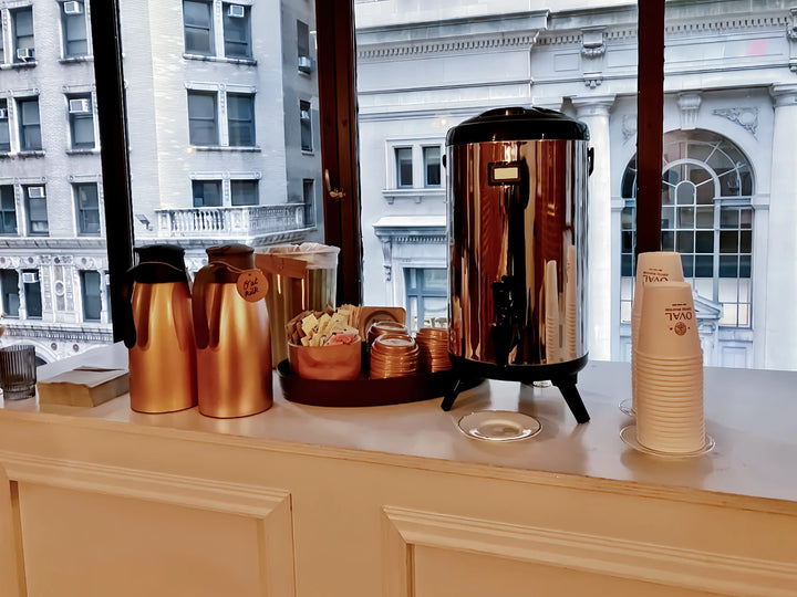 Oval Coffee Roasters drip coffee catering station at Loft 39 in New York City, with a stainless-steel coffee urn, gold thermal carafes, condiments and stacked Oval cups beside large windows.