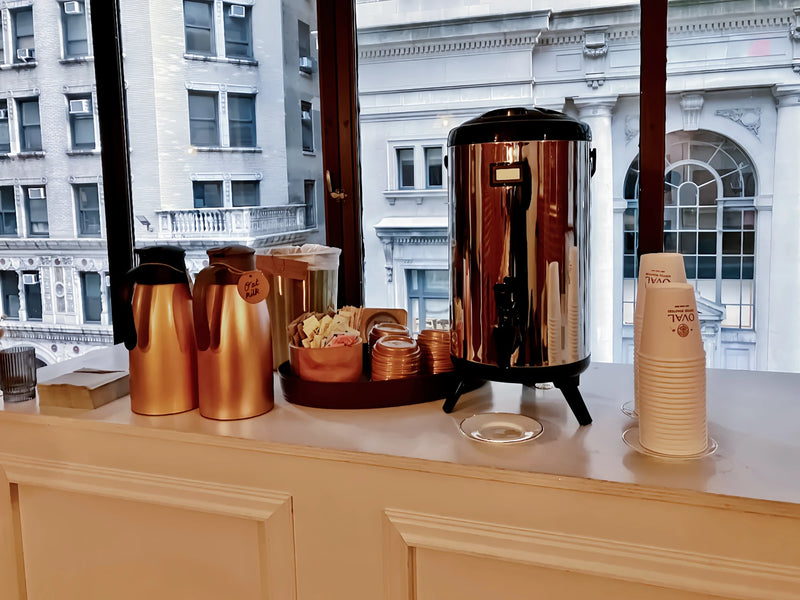 Oval Coffee Roasters drip coffee catering station at Loft 39 in New York City, with a stainless-steel coffee urn, gold thermal carafes, condiments and stacked Oval cups beside large windows.