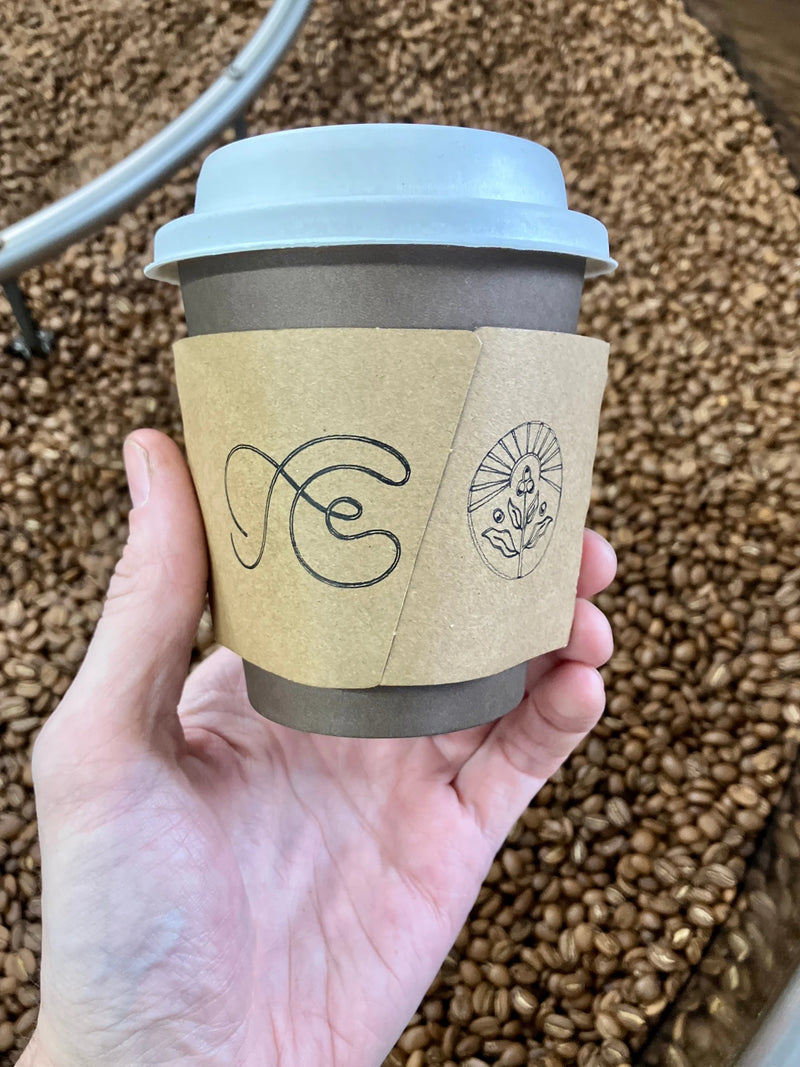A hand holds a paper coffee cup with a pale blue lid and kraft sleeve stamped with Fax Copy Express and Oval Coffee Roasters logos, above roasted beans in a cooling tray at Komune in New York City.