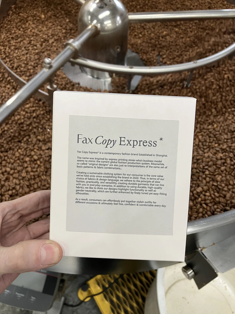 A hand holds a cream-colored Fax Copy Express custom coffee box above a roasting machine’s cooling tray filled with beans, from Oval Coffee Roasters’ light-roast natural-process coffee collaboration.