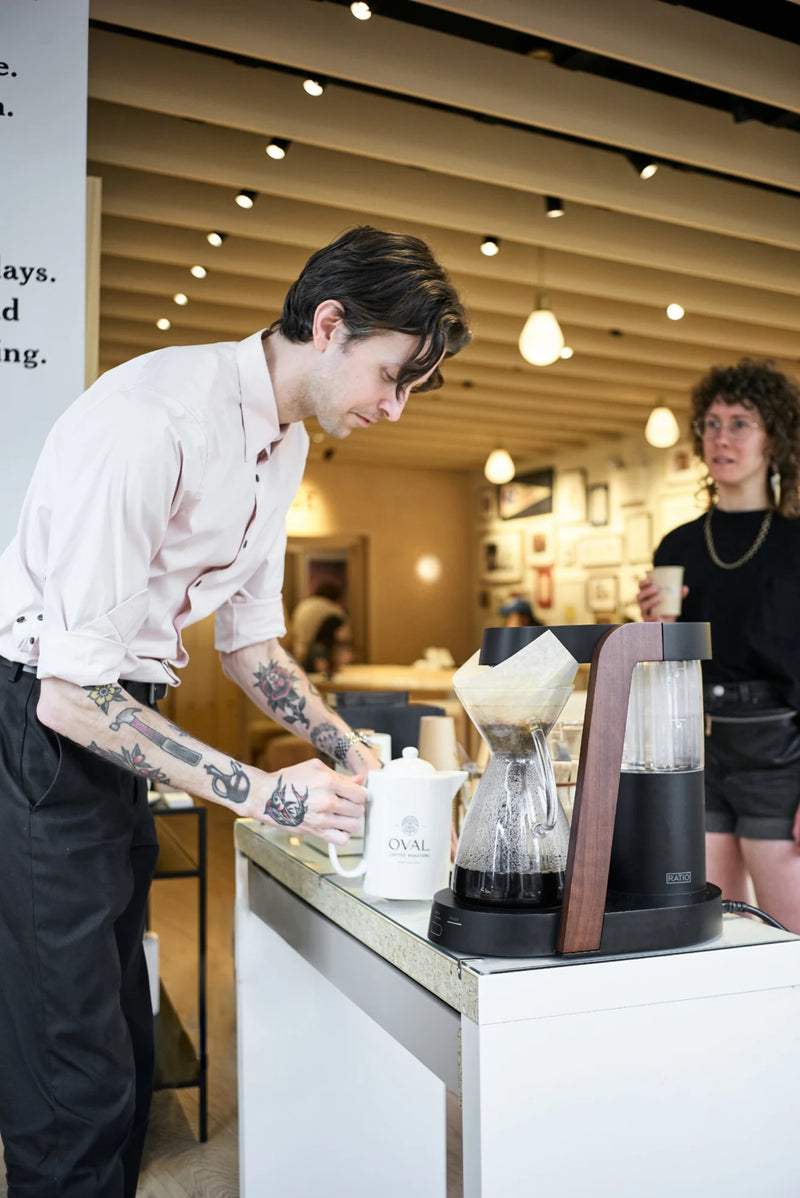 Christopher Alspach preparing Oval coffee beside a wood-accented Ratio Eight brewer and an Oval-branded white pot at Framebridge in Greenwich Village, New York City, while a guest holds a cup nearby.