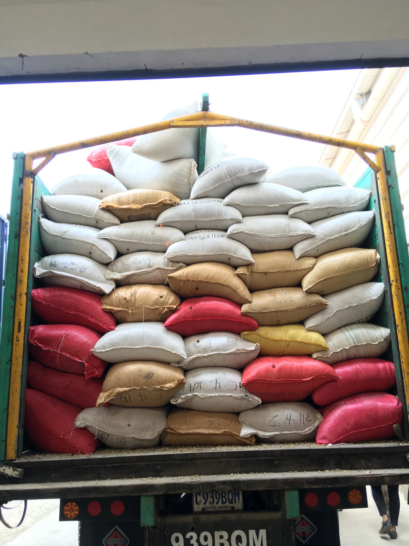 Red, white and beige coffee sacks stacked high in the open bed of a green truck at a coffee mill in Guatemala City, ready for transport.