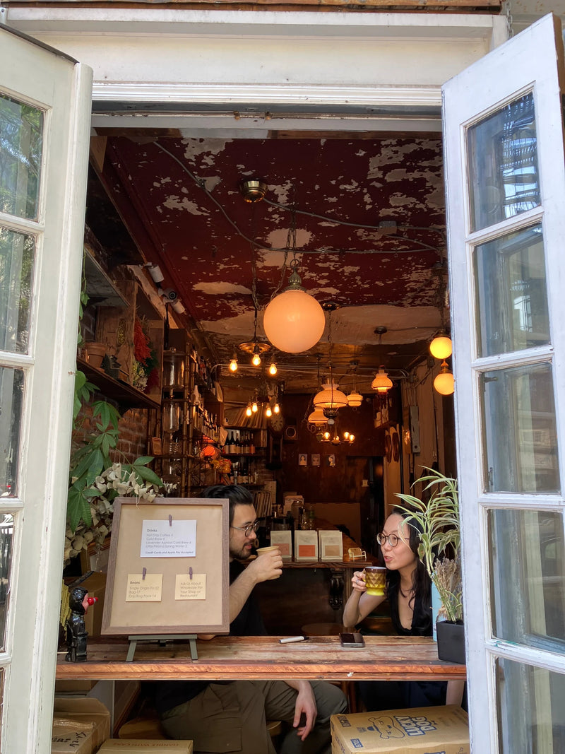 View through an open white-framed window at Oval’s Hard To Explain East Village pop-up, with two guests drinking coffee beside a framed menu, potted plants and warm pendant lights.