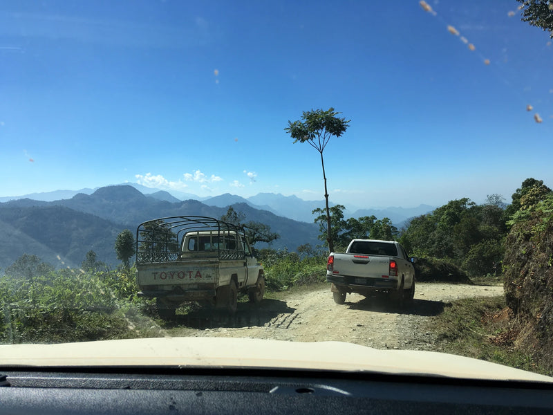 View through a vehicle windshield of two pickup trucks on a winding dirt road near a coffee cooperative in Huehuetenango, Guatemala, with layered mountain ranges beneath a blue sky.