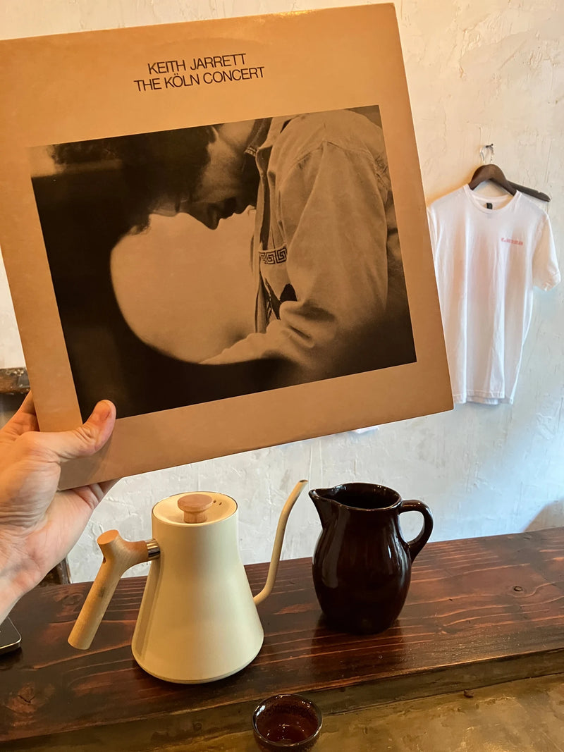 A hand holds Keith Jarrett’s The Köln Concert record sleeve above a cream Fellow Stagg gooseneck kettle and dark brown vintage redware carafe on the wooden counter at an Oval coffee pop-up.