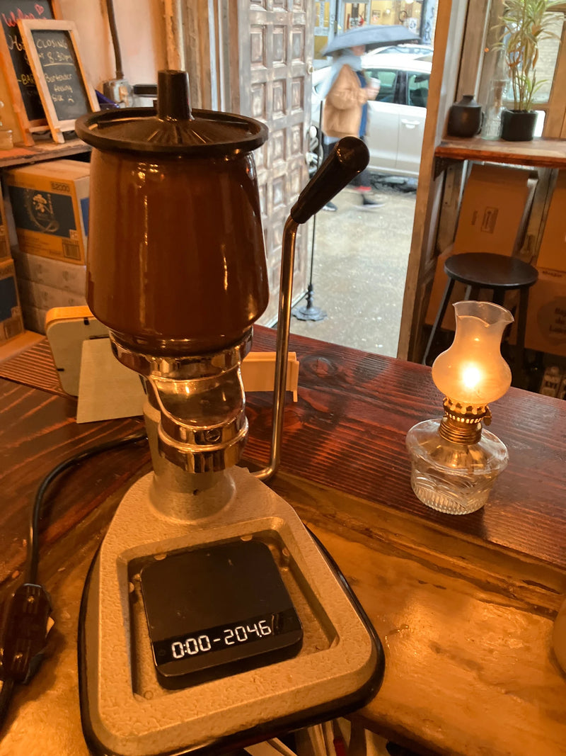 Vintage brown La Peppina lever espresso machine with a digital coffee scale beside a glowing glass oil lamp on a wooden counter at Oval’s Hard To Explain pop-up in New York City’s East Village.