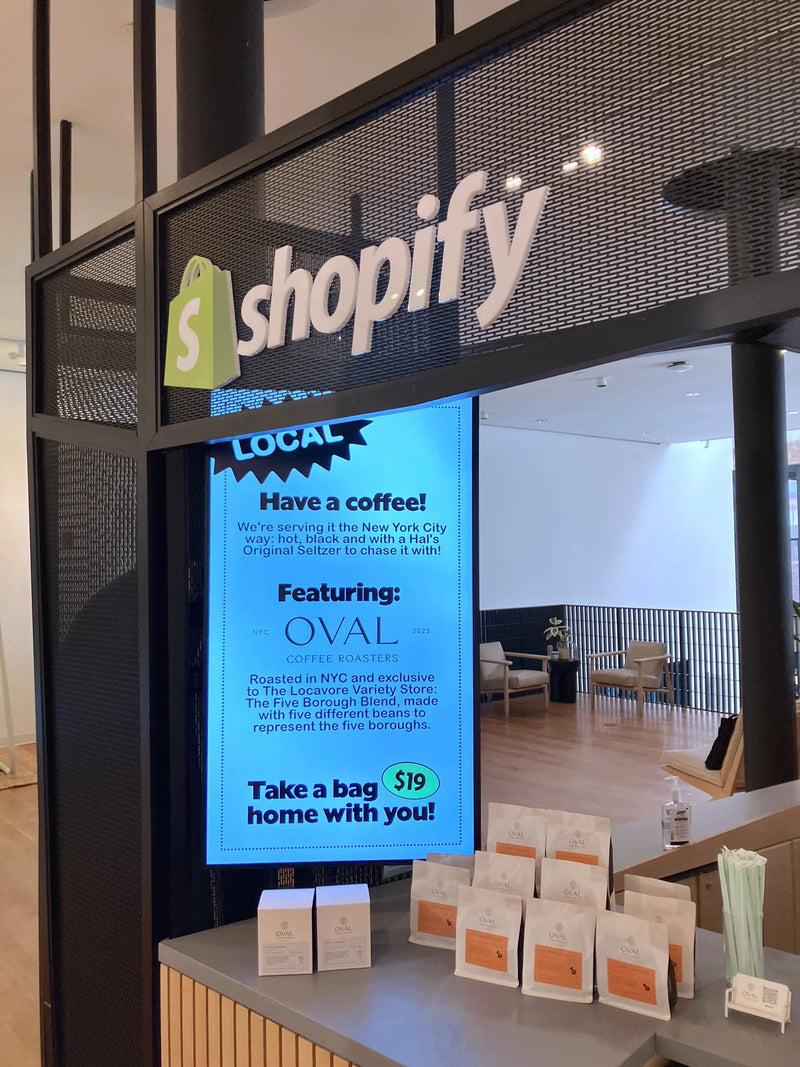 The Locavore custom Oval Coffee Roasters coffee bags and pour-over boxes displayed beneath Shopify signage at its flagship office, beside a screen introducing the Five Borough Blend.