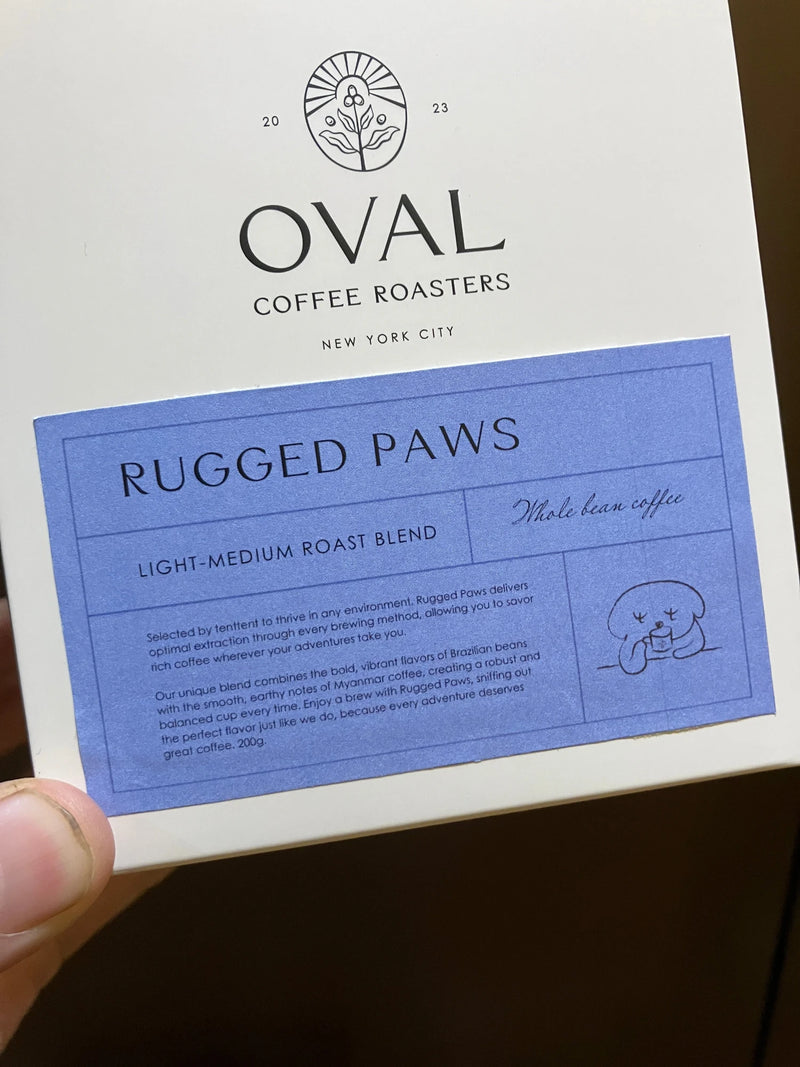 Close-up of a hand-held Oval Coffee Roasters Rugged Paws package created for Tentent, with a lavender label identifying light-medium roast whole-bean coffee and a small dog illustration.
