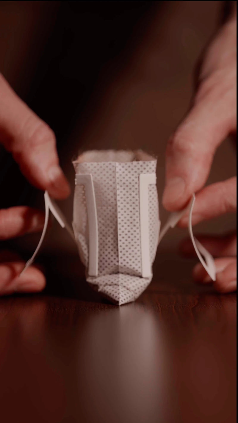 Hands unfold the paper support arms on both sides of an Oval Coffee Roasters single-serve pour-over drip bag on a wooden surface, preparing the filter to hang over a cup for brewing.