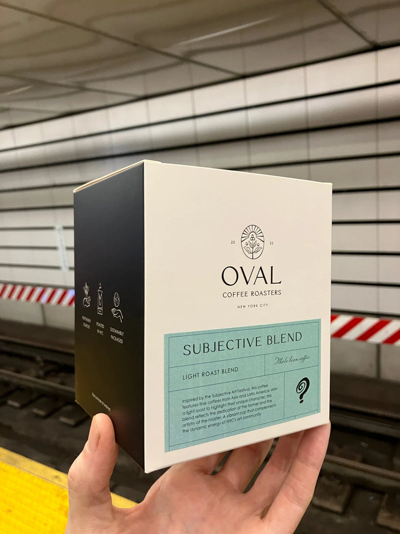 A hand holds Oval Coffee Roasters Subjective Blend custom coffee packaging for Aviva Jifei Xue’s Subjective Arts Festival on a New York City subway platform, with a mint-green light-roast label and black side panel.
