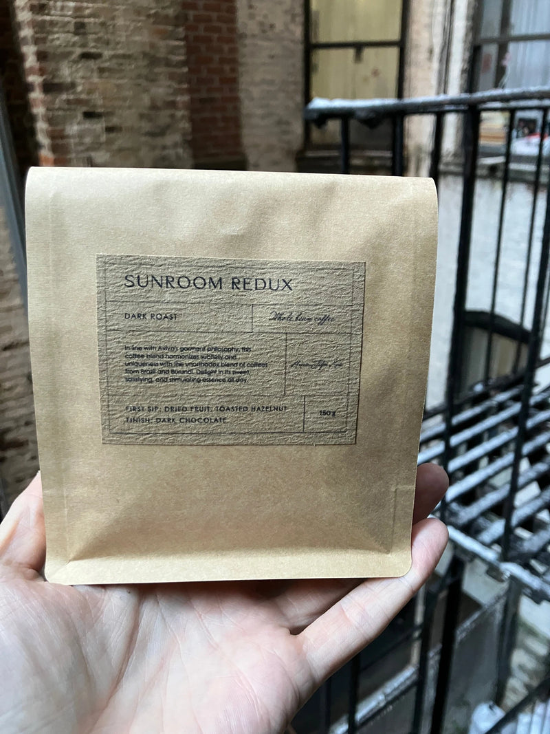 A hand holds a kraft pouch of Sunroom Redux custom whole-bean coffee for Aviva Jifei Xue, with a textured gray dark-roast label in front of a studio window, brick wall and fire escape.